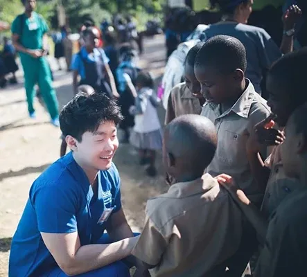 Dr. Lee in blue scrubs kneeling and smiling while interacting with children outdoors during a dental mission trip