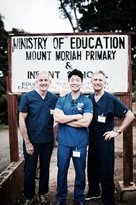 Dr. Lee and two fellow dental professionals in blue scrubs posing in front of a school sign during a mission trip